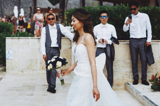 Cheerful Asian bride walks before groomsmen outside