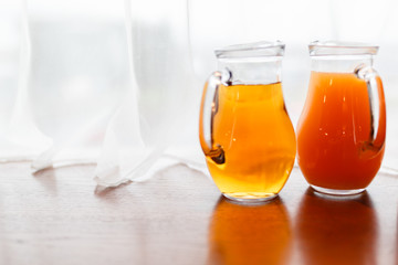 Two jugs with summer juice amid curtains of white cloth. A pitcher of orange and peach juice on the window sill
