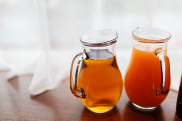 Two jugs with summer juice amid curtains of white cloth. A pitcher of orange and peach juice on the window sill