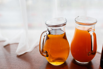 Two jugs with summer juice amid curtains of white cloth. A pitcher of orange and peach juice on the window sill