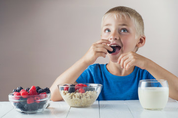 Happy, smiling child eating oatmeal with berries. The concept of a healthy breakfast for children, porridge with raspberries and blackberries, a glass of yogurt on white wooden table.