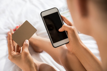 Young woman using smartphone for shopping online with credit card in bedroom