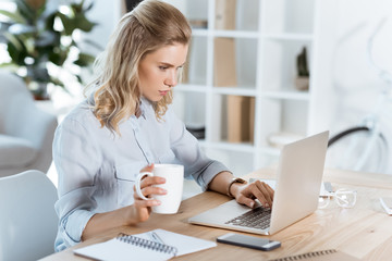 side view of young businesswoman with cup of coffee in hand typing on laptop in office