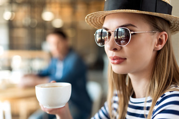portrait of stylish woman in hat and sunglasses holding cup of coffee in cafe