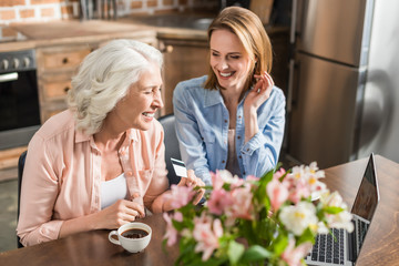 young and senior women sitting at table and using laptop in kitchen