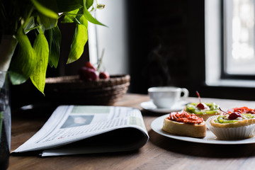 Still life of newspaper, breakfast with cakes and hot coffee on kitchen table in front of window