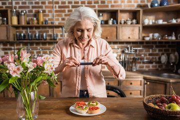 Portrait of senior woman photographing plate with food at kitchen