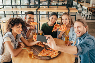 Young man taking selfie with multiethnic friends having pizza in cafe