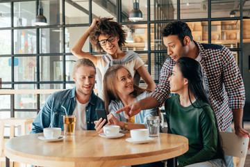 Young happy multiethnic friends looking at smartphone while resting in cafe