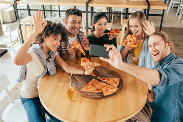 Young man taking selfie with multiethnic friends having pizza in cafe
