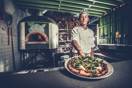 Food concept. Preparing traditional italian pizza. Young smiling chef in white uniform and gray hat show ready dish with green rucola herbs in interior of modern restaurant kitchen. Ready to eat.