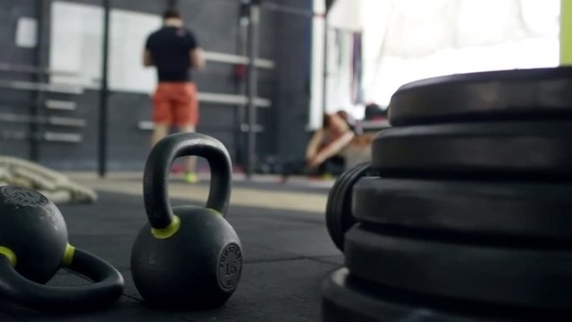 PAN of kettlebells and weight lying on mat floor of gym; male athlete standing in background 