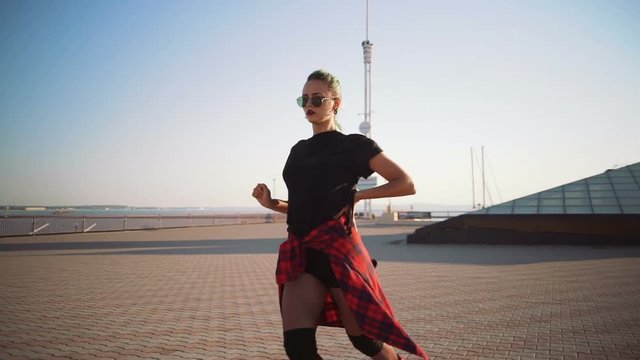 Young stylish funky girl with green hair riding roller skates and dancing near sea port during sunset, slow motion
