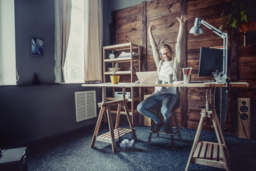 Freelancer man at his home office. Young man working online streching hands up.