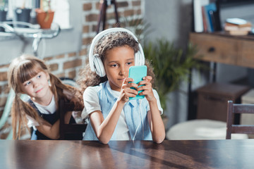 little girl looking how african american friend in headphones using smartphone while siting at table