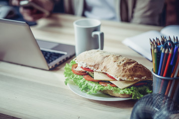 Naklejka premium Close up view of working palce of freelancer during lunch. Wooden desk with laptop, cup of coffee and sandwich on it.