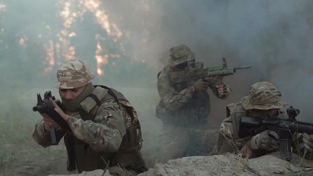 Men in military uniform with guns sitting behind rock shooting and fighting. A bomb explodes nearby and soldiers fall into the trench.