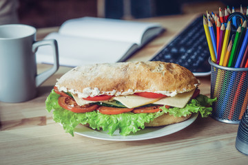Close up view of sandwich on table in frelancer home office. Wooden desk, sandwich on front view, notebook, keyboard and cup on backdrop.