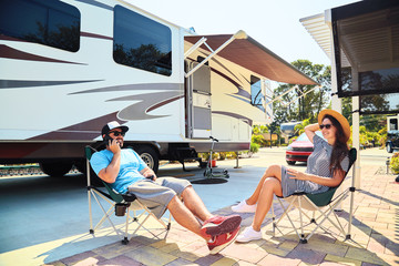 Young couple sits near camping trailer,smiling.Men talks on mobile phone and uses electronic...