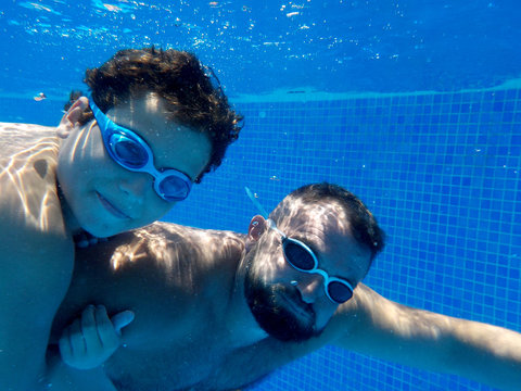 Son And Dad With Goggles Diving Together In A Swimming Pool..