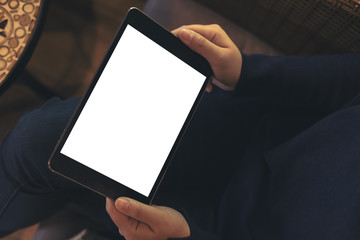 Mockup image of woman's hands holding black tablet pc with blank white screen on thigh with wooden floor background in modern cafe