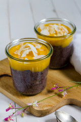 Black rice pudding with mango and coconut flakes in the glass jar.  White background, wooden table.