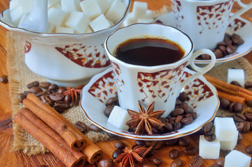 A cup of black coffee with sugar bowl among the sackcloth, cinnamon bark, whole grains of coffee, anise stars and lump sugar on wooden table