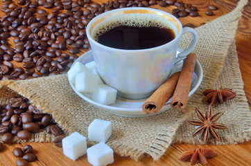 A cup of black coffee with cinnamon bark and lump sugar among the sackcloth, whole grains of coffee and anise star