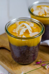 
Black rice pudding with mango and coconut flakes in the glass jar.  White background, wooden table.
