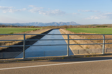 Canal de agua para el riego en paisaje agricola, montañas al fondo y camino en paralelo al canal....