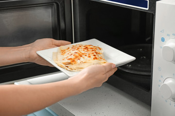 Woman putting plate of lasagna into microwave oven in kitchen