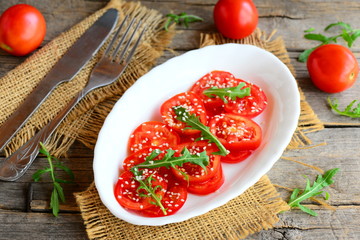 Tomato, arugula salad. Tasty and diet salad with tomatoes, arugula and sesame seeds on a white plate. Fork, knife, burlap textile on a vintage wooden table. Rustic style