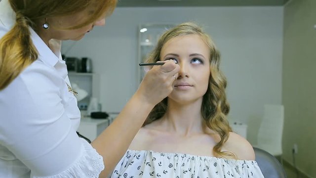 Professional makeup artist applying eye pencil to blond models eyelid for doing eyelid arrows. Front view. Young attractive girl with long curly blond hair and blue eyes.