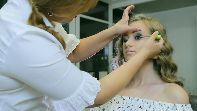 Professional makeup artist applying mascara to blond models eyelashes. / Professional makeup artist applying mascara models eyelashes. Young attractive girl with long curly blond hair and blue eyes.