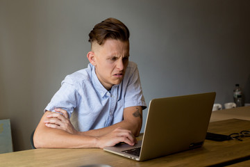 Disgusted and overwhelmed young man with trendy haircut stares at screen of his laptop and social media information, annoyed and surprised by data