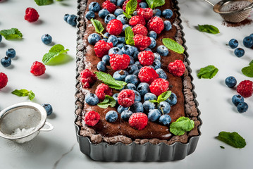 Summer homemade baked pastry. Chocolate cake tart with chocolate cream, fresh raw berries blueberry raspberry, decorated with mint leaves, powdered sugar. On white marble table, copy space