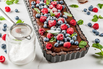 Summer homemade baked pastry. Chocolate cake tart with chocolate cream, fresh raw berries blueberry raspberry, decorated with mint leaves, powdered sugar. On white marble table, copy space close view