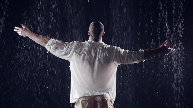 view from the back. average plan. a large man in white clothes standing under a large rain threw his arms to the sky and pray to God