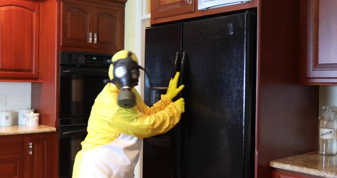 Woman in yellow Haz Mat gear with face mask inside home looking inside refrigerator.