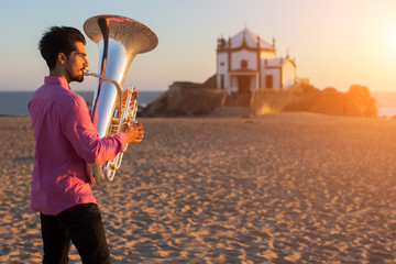 Young man playing the trumpet on the sea coast. Tuba instrument. In the background is the Chapel Senhor da Pedra at Miramar Beach, Atlantic ocean, Porto, Portugal..