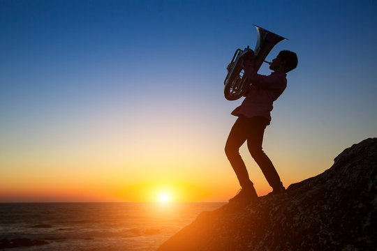 Silhouette of musician playing the trumpet on rocky sea coast during sunset. Tuba instrument.