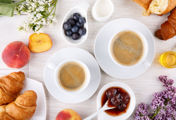 Breakfast - two cups of coffee, croissants, jam, honey and fruits on white table.