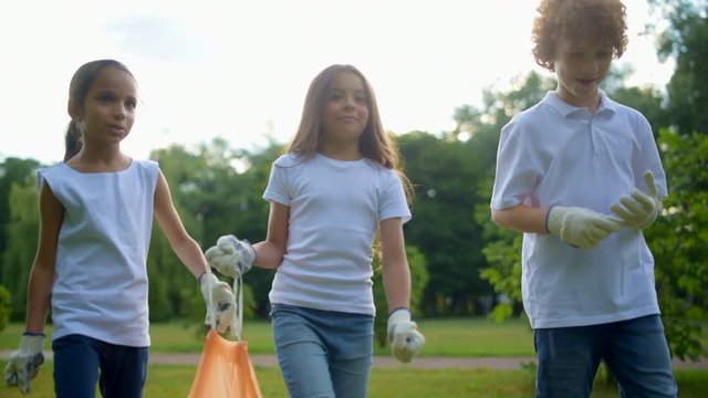 Cheerful little volunteers holding garbage bag and walking around park