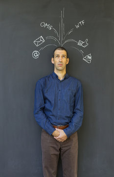 Single white adult man standing in front of a blackboard with drawn social media icons symbolizing frustration, stress and overload from modern communication