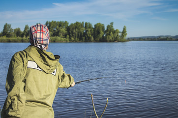 A young woman in green clothes is standing on the river bank and fishing with a fishing rod.