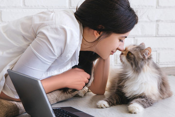 Young woman and her lovely cat rubbing noses each other. Fluffy family pet lays on working table with laptop nearby
