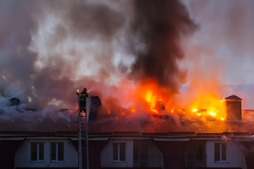 Burning fire flame with smoke on the apartment house roof in the city, firefighter or fireman on the ladder extinguishes fire