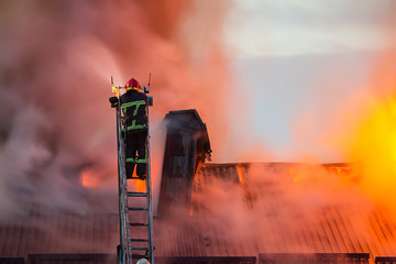 Firefighter or fireman on the ladder extinguishes burning fire flame with smoke on the apartment house roof © O.Farion