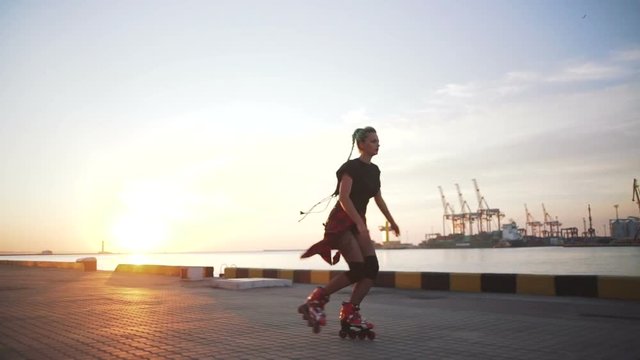 Young stylish funky girl with green hair riding roller skates and dancing near sea port during sunset