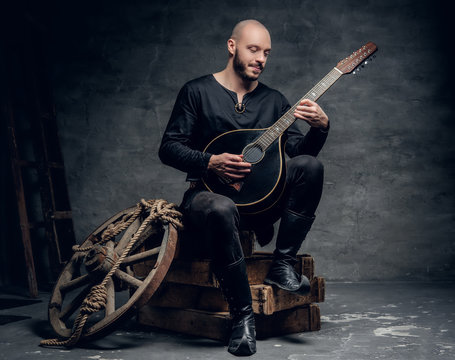 Traditional folk musician dressed in vintage Celtic clothes sits on a wooden box and plays mandolin.
